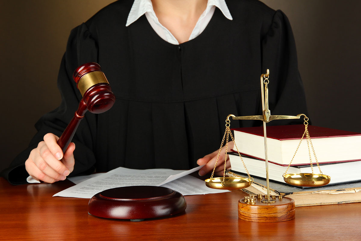 A judge in a black robe is seated at a wooden desk, holding a gavel in one hand and striking it on a wooden sound block. In the foreground, there is a set of golden scales of justice, and beside it are several large, thick books with red covers, symbolizing law and legal texts. The judge is focused on documents on the desk, indicating they are in the process of delivering a verdict or making a legal decision. The background is plain and dark, keeping the focus on the judicial elements and the judge.